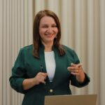 Confident woman in a green blazer giving a presentation in a modern office setting.